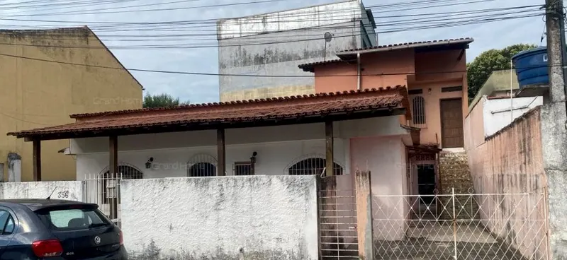 Fachada da casa com varanda de telhas, muro e portões, e construção de dois andares ao fundo